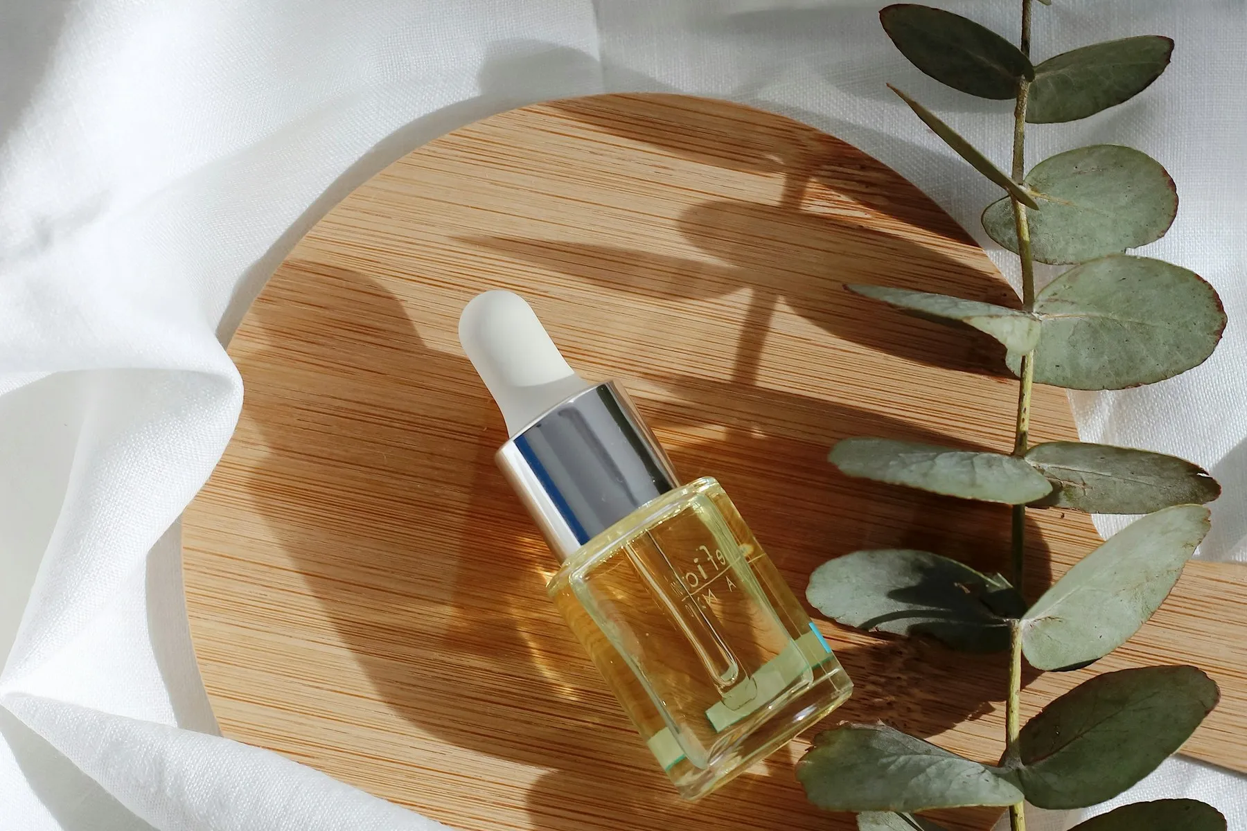 A glass dropper bottle on a wooden tray with eucalyptus and white linen