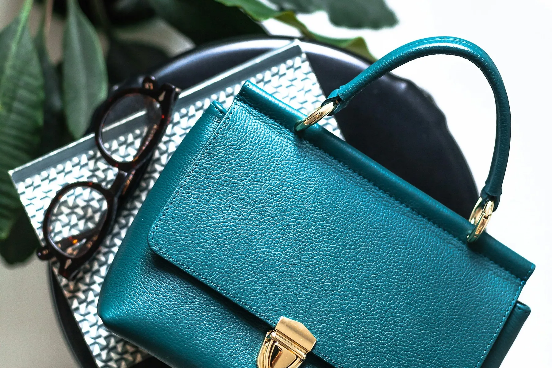 A teal leather top-handle bag resting on books beside a pair of glasses