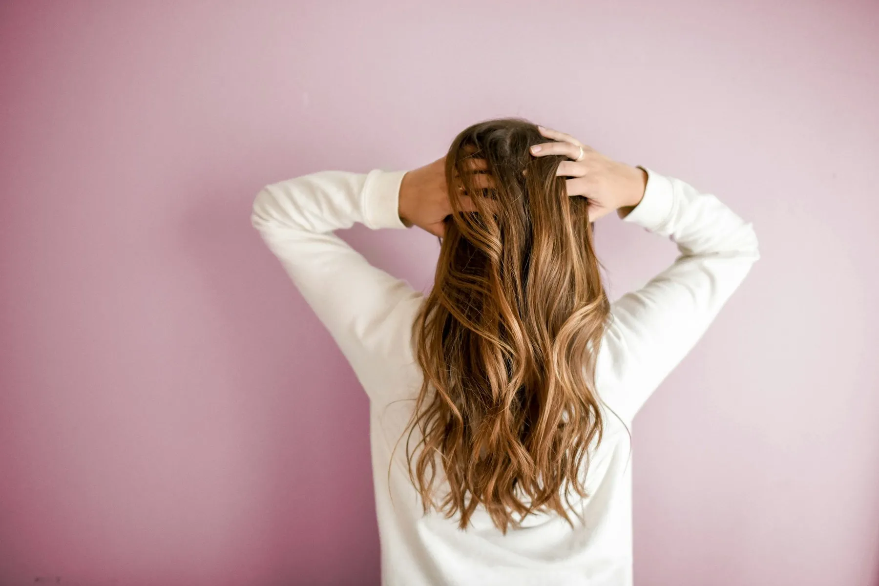 A woman with long, wavy brown hair facing a pink wall